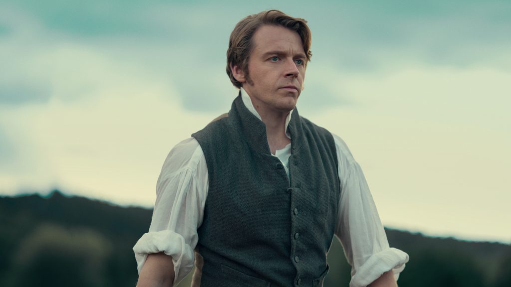 "A man in historical clothing stands outdoors with a serious expression, wearing a vest over a white shirt, with hills and trees in the blurred background under a cloudy sky."