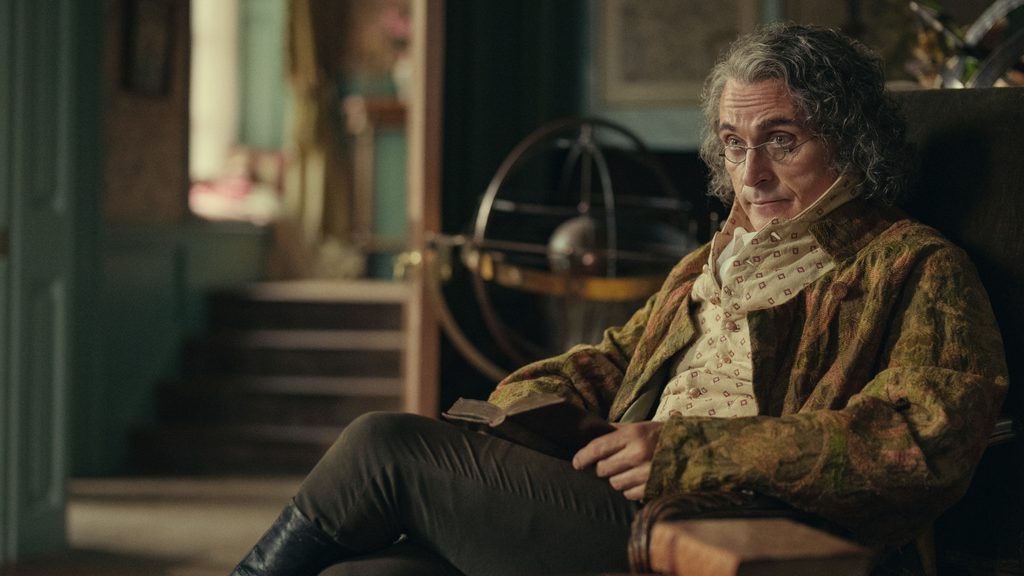"Older man with curly gray hair and glasses sits in an ornate, vintage room holding a book, surrounded by antique furnishings, with an armillary sphere and staircase in the background, evoking a historical or scholarly atmosphere."