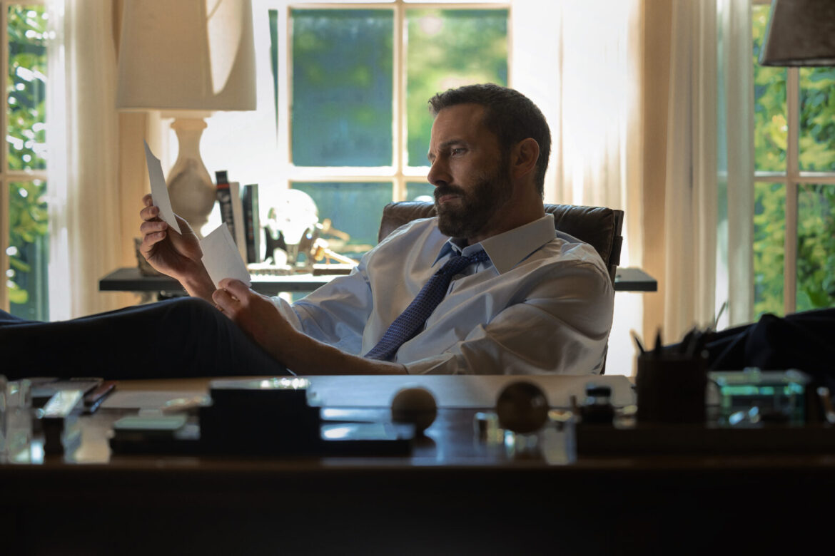 "Man in dress shirt and tie sits at a desk in a sunlit office, reading papers with feet up. Desk has pens, papers, and lamps. Large windows and trees outside create a bright, contemplative setting."
